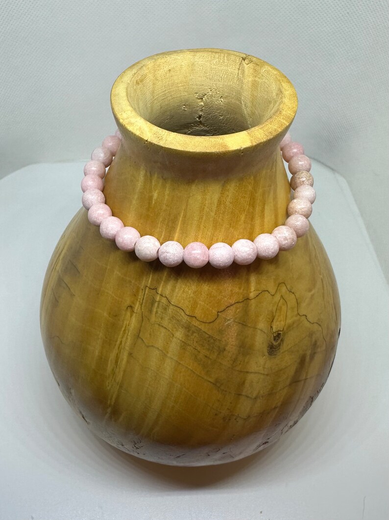 K&ouml;nnte beinhalten: Ein Armband aus rosafarbenen Edelsteinen mit runden Perlen auf einer Holzoberfl&auml;che.