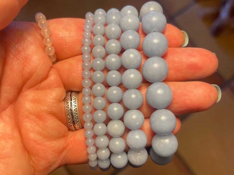 Puede incluir: Un conjunto de seis pulseras hechas con cuentas de piedras preciosas de color azul claro. Las pulseras est&aacute;n apiladas una encima de la otra y las est&aacute; sujetando una mano.