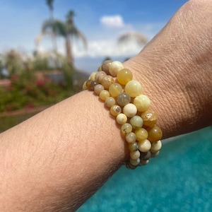 Könnte beinhalten: Drei gestapelte Armbänder mit Perlen. Die Armbänder haben runde Perlen in verschiedenen Größen und Farbtönen von Gelb, Creme und Braun. Die Perlen scheinen aus Stein zu sein. Die Armbänder werden am Handgelenk getragen.