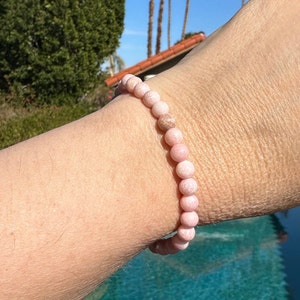 K&ouml;nnte beinhalten: Ein Armband aus rosafarbenen Edelsteinen mit runden Perlen. Das Armband wird am Handgelenk getragen.
