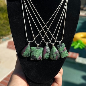 May include: Five green and pink gemstone pendants on silver chains. The pendants are in various shapes, including teardrop, square, and rectangular.
