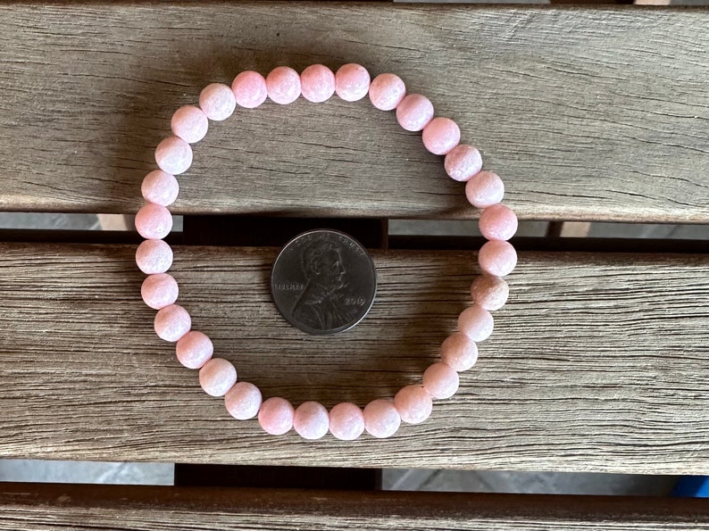 K&ouml;nnte beinhalten: Ein Armband aus rosafarbenen Edelsteinen mit runden Perlen. Das Armband liegt auf einer Holzoberfl&auml;che neben einem deutschen Centst&uuml;ck zum Gr&ouml;&szlig;envergleich.
