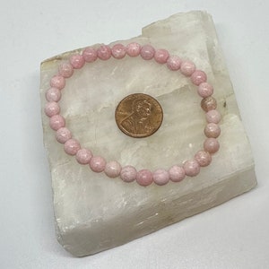 K&ouml;nnte beinhalten: Ein Armband aus rosafarbenen Edelsteinen mit runden Perlen. Das Armband liegt auf einer wei&szlig;en Oberfl&auml;che neben einem US-Cent.