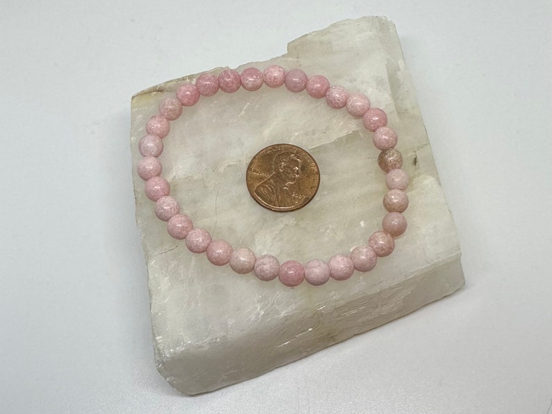 K&ouml;nnte beinhalten: Ein Armband aus rosafarbenen Edelsteinen mit runden Perlen. Das Armband liegt auf einer wei&szlig;en Oberfl&auml;che neben einem US-Cent.