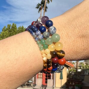 May include: A bracelet made of colorful gemstone beads. The bracelet is layered with different colors of beads, including clear, purple, blue, green, yellow, brown, red, and orange. The bracelet is worn on a person's wrist.