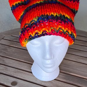 Puede incluir: Un gorro de punto colorido con un patrón de rayas en tonos de rojo, naranja, amarillo, verde, azul y morado. El gorro tiene una forma única con una parte superior puntiaguda y un ala ancha.