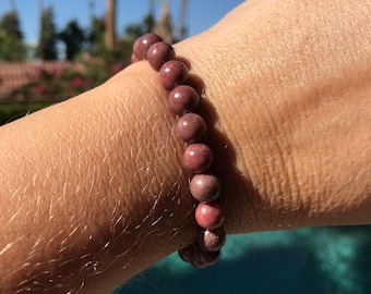 May include: A beaded bracelet made of round, pinkish-brown stones. The bracelet is worn on a wrist, with a blurred background of a pool and palm trees under a bright blue sky. The stones appear to be approximately 0.5 inches in diameter.