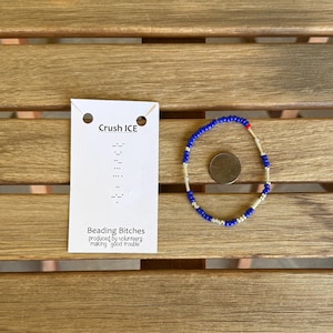 Può includere: Un braccialetto di perline fatto a mano con perline blu e argento, accentuato da una singola perlina rossa. Un'etichetta bianca con il testo "Crush ICE" e "Beading Bitches" è accanto al braccialetto. Il braccialetto ha una circonferenza di circa 18 cm.