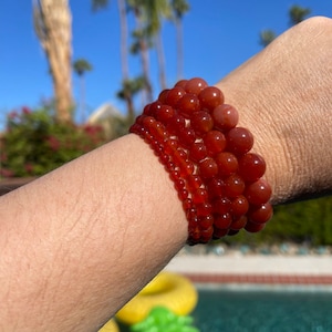 May include: A stack of carnelian bracelets in various sizes, with the largest bracelet at the top and the smallest at the bottom. The bracelets are made of smooth, round beads and are a deep orange-red color.
