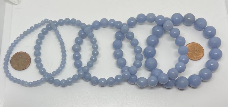 Puede incluir: Cinco pulseras de piedras preciosas de color azul claro de diferentes tama&ntilde;os, dispuestas sobre una superficie blanca. Una moneda de un centavo de d&oacute;lar estadounidense se coloca junto a la pulsera m&aacute;s peque&ntilde;a para comparar el tama&ntilde;o.