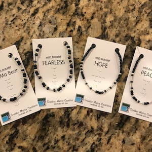 Puede incluir: Cuatro pulseras de cuentas negras y plateadas con tarjetas que dicen "pulsera de deseos" y las palabras "Mama Bear", "Fearless", "Hope" y "Peace" en texto negro. Las tarjetas también incluyen el alfabeto braille para cada palabra.