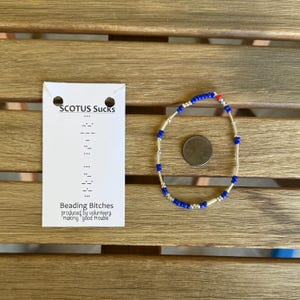 Può includere: Un braccialetto di perline blu e argento, con una singola perlina rossa. Una carta bianca con il testo "SCOTUS Sucks" e "Beading Bitches" è accanto al braccialetto. Il braccialetto è su una superficie di legno.