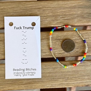 May include: A beaded bracelet with rainbow-colored beads and silver accents. A white card with the text "Fuck Trump" and "Beading Bitches" is next to the bracelet. The card also includes text in braille and the phrase "making good trouble."