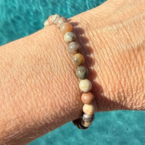 Peut inclure: Un bracelet de perles composé de pierres rondes et lisses dans des tons terreux variés, incluant des nuances de beige, de gris, de brun clair et de brun sourd. Les perles sont enfilées sur un seul rang, montrant des variations naturelles.