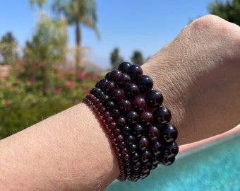 May include: A stack of six garnet bracelets, each with a different size of beads, worn on a wrist against a blue background.