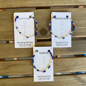 Pode incluir: Três pulseiras de contas azuis, prateadas e vermelhas são exibidas em cartões brancos. Cada cartão tem uma frase diferente: "Fuck Trump", "SCOTUS Sucks" e "Crush ICE". Os cartões também incluem o texto "Beading Bitches" e "produced by volunteers making good trouble".