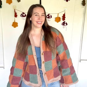 May include: A long-sleeved crochet cardigan featuring a checkered pattern in muted sage green, burnt orange, and dusty rose. The garment has a beige ribbed trim along the front opening and three brown buttons. It is worn over a blue tank top and jeans.