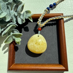 May include: A necklace with a pale yellow, oval pendant with a distinctive pattern. The pendant hangs from a braided cord with blue and red beads. The necklace is displayed on a dark grey background within a wooden frame.