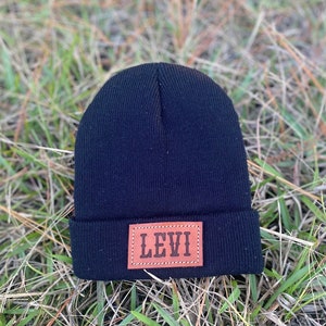 May include: A black beanie hat with a brown leather patch. The patch is rectangular and features the name "LEVI" in capital letters. The hat is made of a ribbed knit material and is resting on a bed of green and brown grass.