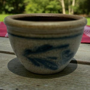 Può includere: Una piccola ciotola in ceramica fatta a mano, nei toni dell'azzurro e del beige. La ciotola presenta un motivo a foglie blu scuro e una forma arrotondata. È appoggiata su un tavolo di legno. Diametro: circa 10 cm.