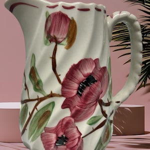 May include: White ceramic pitcher with a twisted body and a handle. The pitcher is decorated with large, red poppy flowers, green leaves, and brown stems. The rim has a scalloped edge with red accents. A decorative floral pitcher.