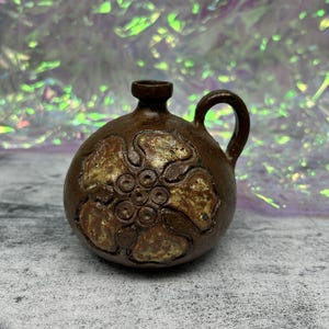 May include: A small, brown ceramic jug with a handle and a decorative floral design. The jug has a rounded body and a small opening at the top. The floral design is in a lighter brown color, creating a contrast against the darker brown background.