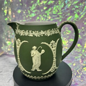 May include: A matte dark green ceramic creamer pitcher featuring white relief ornamentation. The exterior displays a central figure holding a staff and mask, framed by vertical scrollwork and a decorative ivy vine border along the top rim.
