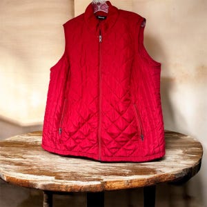 May include: A quilted red vest with a full-length zipper and two side pockets. The vest has a stand-up collar and a diamond pattern throughout. The vest is displayed on a round wooden table.