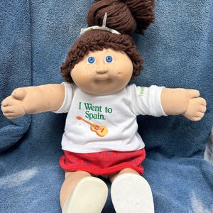 以下が含まれることがあります： 茶色の髪をポニーテールにしたキャベツパッチキッズのドール。白いTシャツに「I Went to Spain.」の文字、赤いスカートを着用しています。青い目とそばかすがあります。
