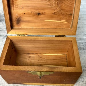 Può includere: Una scatola di legno con un coperchio incernierato e una chiusura in ottone. La scatola è aperta e l'interno è vuoto.