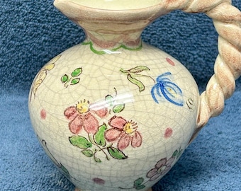 May include: A small, white ceramic pitcher with a twisted rope handle. The pitcher is decorated with a floral pattern in shades of pink, yellow, green, and blue.