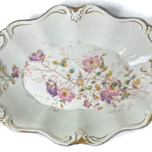 Könnte beinhalten: Eine weiße Porzellanservierschale mit goldenen Verzierungen und einem Blumendesign. Die Schale hat zwei Griffe und ist oval.