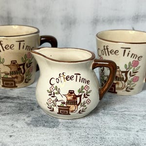 May include: Three ceramic mugs with a brown and white floral design. The mugs feature the text "Coffee Time" and are decorated with a coffee pot and grinder.