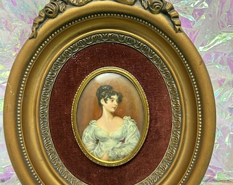 Puede incluir: Un retrato antiguo de una mujer en un marco ovalado dorado. El retrato está sobre un fondo de terciopelo burdeos. El marco tiene detalles ornamentados y una parte superior decorativa. La mujer del retrato lleva un vestido azul claro.