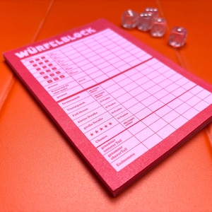 May include: A pink score pad for a dice game with the text W&Uuml;RFELBLOCK printed at the top, featuring a grid for tracking points and five clear, transparent dice positioned near the top right corner of the paper pad.