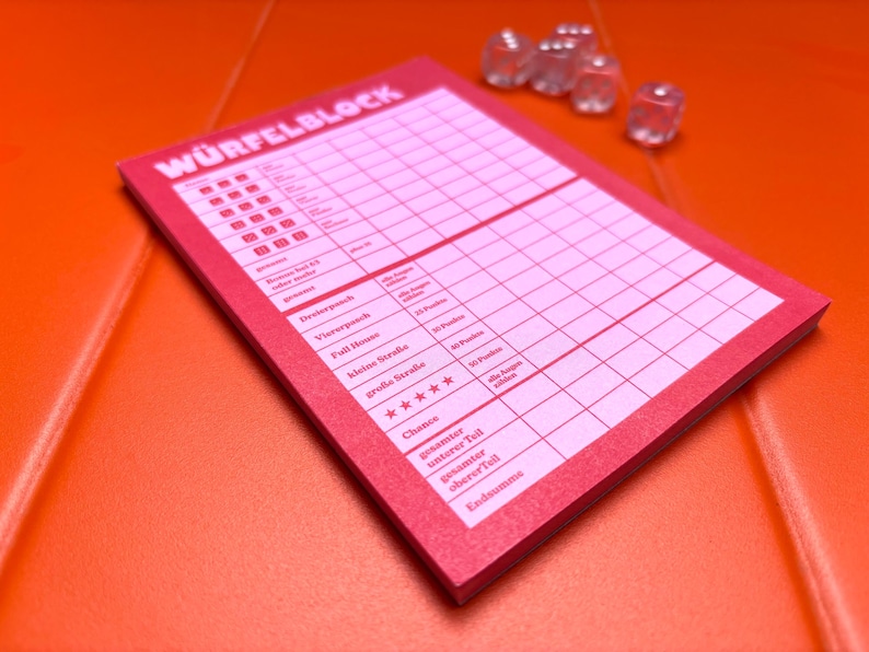 May include: A pink score pad for a dice game with the text W&Uuml;RFELBLOCK printed at the top, featuring a grid for tracking points and five clear, transparent dice positioned near the top right corner of the paper pad.