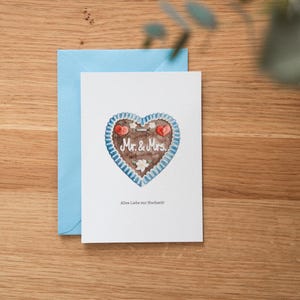 May include: A white greeting card with a watercolor illustration of a heart-shaped gingerbread cookie with the words "Mr. & Mrs." and a blue envelope on a wooden surface. The card has the text "Alles Liebe zur Hochzeit!"