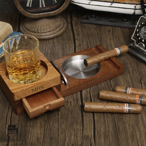 Puede incluir: Un soporte de madera para cigarros y whisky con un caj&oacute;n deslizante. El soporte tiene un cenicero de metal y un vaso para whisky. El soporte est&aacute; personalizado con las iniciales "MHK".