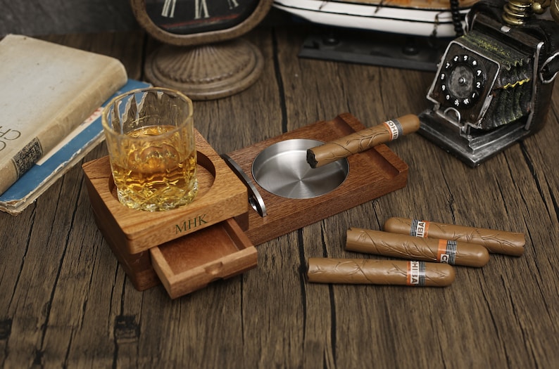 Puede incluir: Un soporte de madera para cigarros y whisky con un caj&oacute;n deslizante. El soporte tiene un cenicero de metal y un vaso para whisky. El soporte est&aacute; personalizado con las iniciales "MHK".