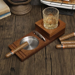 Puede incluir: Porta cigarros y whisky de madera con un vaso de whisky, un cenicero para cigarros y cuatro cigarros.