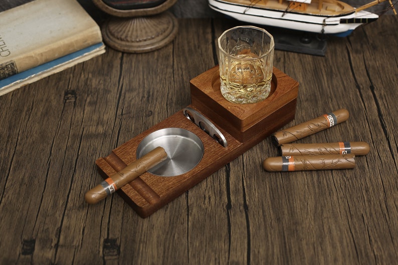 Puede incluir: Porta cigarros y whisky de madera con un vaso de whisky, un cenicero para cigarros y cuatro cigarros.