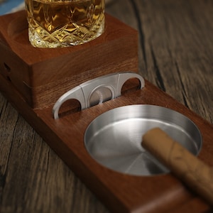 Puede incluir: Un soporte de madera para cigarros y whisky con un cenicero y un cortador de cigarros de acero inoxidable. El soporte tiene un compartimento para un vaso de whisky y una ranura para un cortador de cigarros.