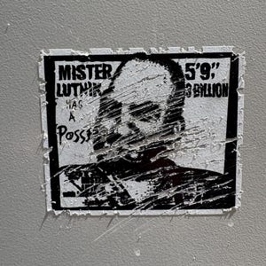 May include: A black and white sticker with the text "Mister Luthik" and "5'9"; $3 Billion" over a portrait. The sticker is affixed to a gray surface and shows signs of wear and tear.