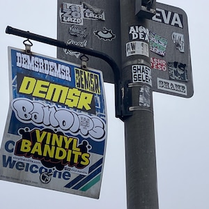 May include: A black and yellow sign with the text "Vinyl Bandits" and a QR code. The sign is attached to a gray pole with several other stickers on it. The stickers include the text "DEMSR", "LAZO", "ASHR ESO", "SHAST SELEK", "SHAME", and "EVA".