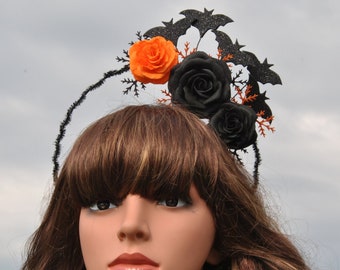 May include: A black headband with a bat design and orange and black fabric flowers. The headband is worn by a person with long brown hair.