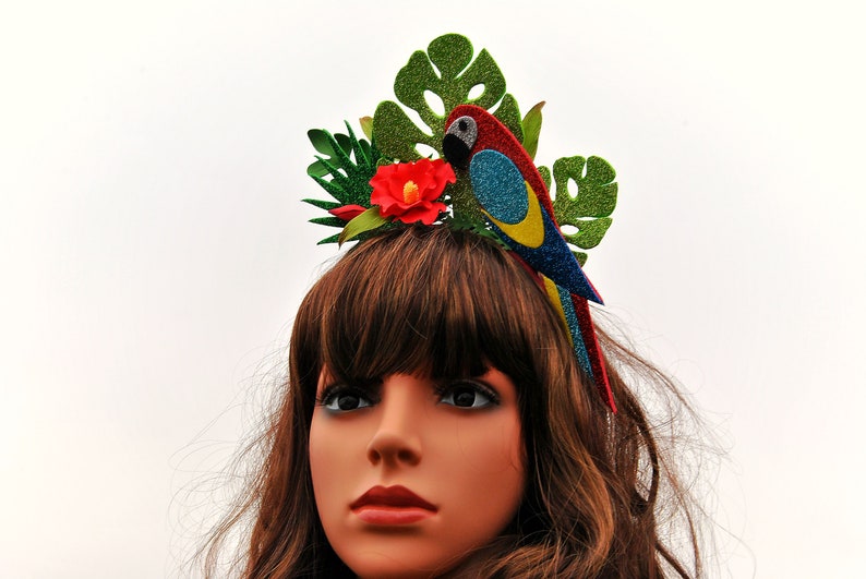 K&ouml;nnte beinhalten: Ein gr&uuml;nes, rotes, blaues und gelbes Glitzer-Papageien-Haarband mit einer roten Blume und gr&uuml;nen Bl&auml;ttern.