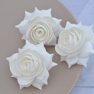 May include: Three white foam roses with a layered, ruffled design. The roses are arranged on a light brown surface.