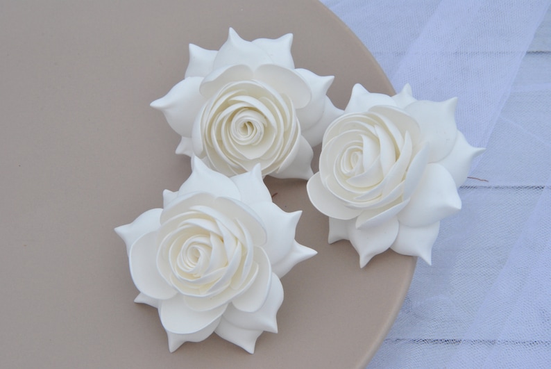 May include: Three white foam roses with a layered, ruffled design. The roses are arranged on a light brown surface.