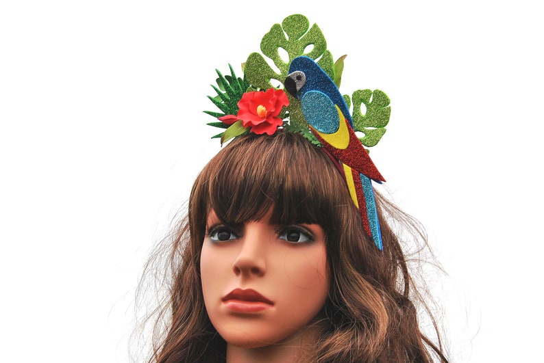 K&ouml;nnte beinhalten: Ein blauer, roter und gelber Glitzer-Papageien-Haarreifen mit gr&uuml;nen Bl&auml;ttern und einer roten Blume. Der Haarreifen wird von einer Mannequin mit braunen Haaren getragen.