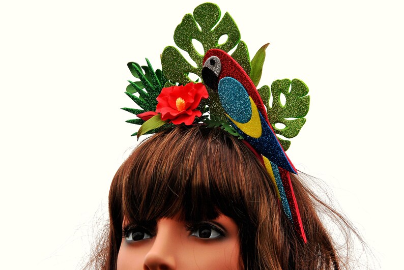 K&ouml;nnte beinhalten: Ein roter, blauer und gelber Papagei mit gr&uuml;nen Glitzerbl&auml;ttern und einem roten Blumenhaarband.